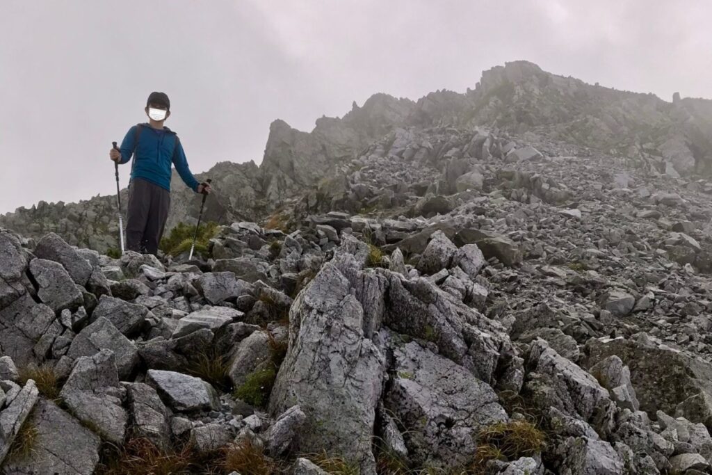 ガスに包まれた岩場でトレッキングポールを持って立つ登山者
