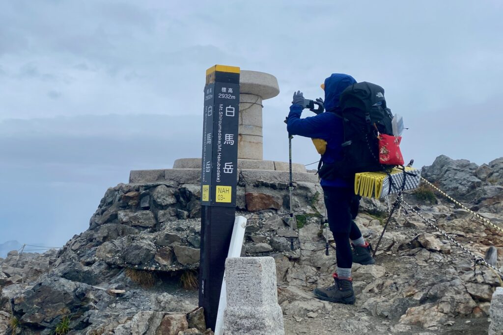白馬岳山頂の標柱の横で写真を撮る登山者